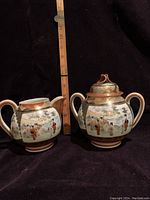 Side view of the creamer and covered sugar bowl with geisha designs and measuring stick for scale