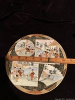 Top-down photo of the round antique Japanese serving plate with a measuring tape showing approximately 8.5 inch diameter, featuring hand painted traditional scenes in multiple panels on a black and gold patterned background.
