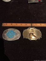 Photo of two belt buckles side by side on black surface next to ruler for size reference