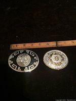 Two collectible belt buckles placed next to a ruler for size reference; left buckle is black enamel with silver etching, right buckle is silver/gold tone with letter D.