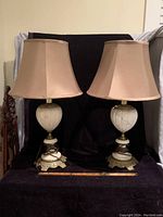 Full front view of both antique brass and alabaster lamps with beige shades together on a table in front of black background.