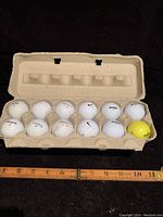 Twelve golf balls in carton, mostly white, one yellow, with visible brands Nike, Nitro, Wilson.