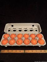 Front view of all 12 orange Titleist golf balls in an open gray carton, arranged in two rows of six each with a measuring scale below showing their approximate size.