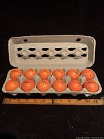 A full view of one dozen orange Titleist golf balls in a gray cardboard carton with a ruler for scale.