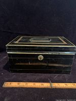 Front view of a black metal cash box with gold trim and central metal handle on the lid, showing keyhole and ruler for size reference.