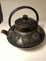 Front view of metal teapot showing arched handle, lid with knob, spout, and embossed medallion patterns on body with dark patina.