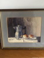 Framed watercolour painting showing a metallic teapot and two crumpets on a plate, on a cloth surface. Painting is behind glass in a brown frame with a gold inner border.