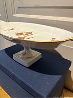 Side view of white pedestal dish showing gold floral design and scalloped oval dish top with gold rim.