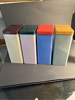 Four rectangular metal containers with different pastel bodies and bright colored lids arranged in a line against a gray backdrop.