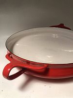 Side angle showing red enamel exterior with handle and white enameled interior of paella dish. Minor surface wear visible.