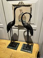 Pair of brass crane bird sculptures displayed on a decorative wooden table with framed vintage art in the background. Shows scale and mounting on green stone bases.