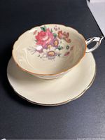 Porcelain teacup and matching saucer shown on dark surface, lighting highlights floral design and gold rim of teacup.