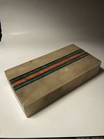 Closed view of rectangular alabaster box with green and pink inlay stripes along the top, showing smooth ivory surface and minor wear.
