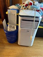 Front view of Arctic King portable air conditioner next to bucket holding window mounting accessories