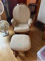 Front view of Dutalier glider rocker chair with matching ottoman, beige striped upholstery, positioned on wood floor near a fan and furniture.