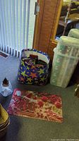 Photo showing Rubbermaid wrapping paper storage container filled with opened rolls, large colorful gift bag containing bags and Christmas wrap accessories, gingerbread men door mat on the floor