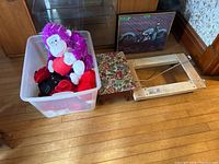 View of plastic bin with Valentine’s Day themed stuffed animals (pink and red gorilla), foam heart bags, cards, and other small decorations alongside floral footstool and wooden dolly frame on floor.