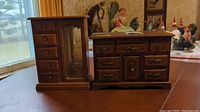 Two wood jewelry boxes resembling a dresser and a wardrobe, placed on a wooden table, with visible landscape background.