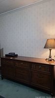 Front view of Malcolm wood dresser with nine drawers, ornate top carvings, metal pulls, placed against wallpapered wall with lamp on top.