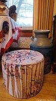 Milk can and velour floral stool side by side near a window with curtains and other household items in background.