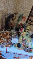 Photo showing a variety of rooster-themed items including large and small figurines, metal jug, woven basket, decorative plates, and several fabric kitchen items like placemats and towels.