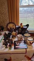 View of the entire set of rooster-themed items on a table with beige tablecloth, showing multiple rooster figurines, mugs, plate, trivet, and candleholders.
