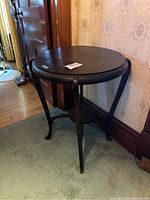Round wooden occasional table seen from a front side angle showing the round top, three legs, and lower shelf against a wooden floor and wall background.