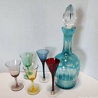 Full set of one blue cut glass decanter with stopper and five colored cocktail glasses arranged in front.