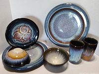 Seven Bill Campbell studio pottery pieces including plates, bowls, and mugs arranged to show overall condition and glaze colors.