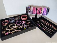 All bracelets and bangles in the lot arranged on a two-tier black display and in black trays, showing variety of styles and colors with dominant purples and pinks.