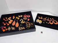 Full overview of two black trays containing multiple pairs of orange and multicolored costume pierced earrings arranged on black velvet underlay, showing variety in size and style.