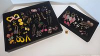 Black tray holding multiple pairs of colorful costume pierced earrings arranged separately showing variety of yellow, pink, purple, and gold toned earrings.