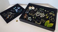 Full view of two black displays holding a variety of green and blue costume pierced earrings including hoops, dangles, studs, and leaf shaped pieces.