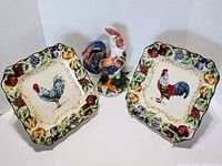 Photo showing all five pieces: four rooster plates with fruit borders and one ceramic rooster statue, arranged together.