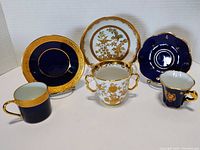 Three tea cup and saucer sets arranged on white surface. Includes deep blue Limoges set, white Limoges two-handled cup set with matching saucer, and navy blue Nestle replica set.