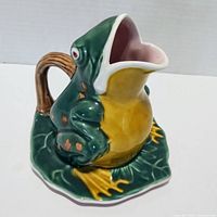 Front-left angled view showing frog pitcher with mouth spout, wrapped arms, and lotus leaf base