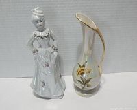 Front view of Victorian lady figurine next to iridescent gold-trimmed pitcher vase with floral decoration.