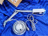 Infrared heat therapy lamp showing circular lamp head, protective grid, white power box, adjustable arm, and power cord with plug on blue fabric background.