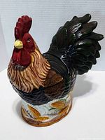 Front and side view of the large ceramic rooster cookie jar showing detailed front feathers, red comb and wattle, body feathers in brown, black, and green, and the base with corn motif.