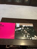 Two Bob Dylan vinyl LP albums on a dark wooden table. One is the album 'A Rare Batch of Little White Wonder' in a bright pink cover, the other is the double album 'Time Out of Mind' with a black and white cover image.