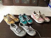 Overview of all four pairs of shoes on brown surface showing colors and style, including yellow Vans, teal Vans with checkered sole, gray Vans, and pink and white running shoes.