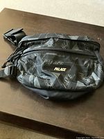 Front view of black Palace fanny pack with all-over logo pattern and front zip pocket.