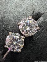 Close-up of one pair of 14k white gold moissanite stud earrings, round brilliant cut stones reflecting light on a black textured background.