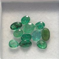 Eleven loose emerald gemstones on white cushioned background, showing various shapes and green shades
