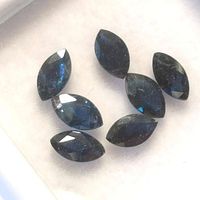 Seven marquise cut dark blue sapphire gemstones, showing facets and natural inclusions, arranged on a white display background.