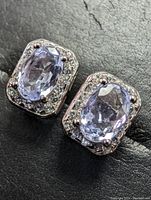 Close-up image of a pair of sterling silver earrings each featuring an oval natural tanzanite stone surrounded by smaller clear stones on a black textured background.