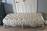Front view of a rectangular white upholstered bench with deep button tufting, scalloped carved wooden legs, and nailhead trim.
