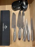 Five Chicago Cutlery knives laid out with an electric knife sharpener and the box for the santoku knife. Knives clean with minor signs of use.