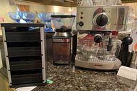 Wide view showing espresso machine, coffee grinder, black plastic storage unit with drawers, milk frothing pitcher, and glass cups on countertop.