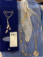 Four necklaces displayed on blue fabric and navy blue display board including silver snake chain with pearl-like drop beads, long gold-plated bead necklace, sheer ivory ribbon necklace with blue charm, and silver chain with round pendant.